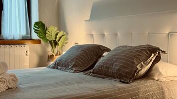 A neatly made bed with a striped bedspread and two pillows, a radiator, a plant, and a window with curtains.