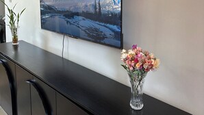 Ein Flachbildfernseher an der Wand, ein dunkles Holzschreibtisch mit Blumen in einer Vase und eine Topfpflanze.