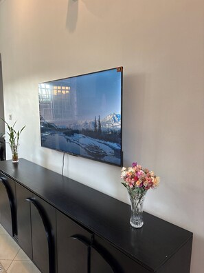 Ein Flachbildfernseher an der Wand, ein dunkles Holzschreibtisch mit Blumen in einer Vase und eine Topfpflanze.