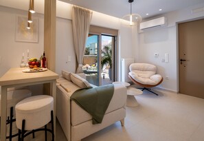 A modern living room with a sofa, a chair, a dining table with fruit, and a view through a glass door. - Casa Doma Luxury Suite with Heated Mini Pool and Private Parking (Rethymno Town)