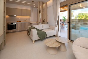 A modern living room with a sofa, two round tables, a kitchen in the background, and a sliding glass door leading to an outdoor area. - Casa Doma Luxury Suite with Heated Mini Pool and Private Parking (Rethymno Town)