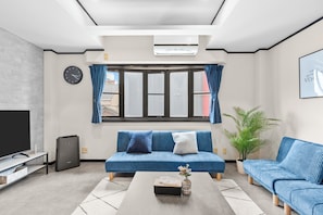 A modern living room with a blue sofa, a coffee table, a TV, and a large window with curtains. - Smart Stay Inn EbisuNishi (Osaka)