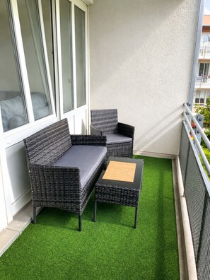 Ein Balkon mit einer schwarzen Rattan-Couch und passendem Hocker, ein kleiner Tisch mit Bambusmatte und einer weißen Wand.