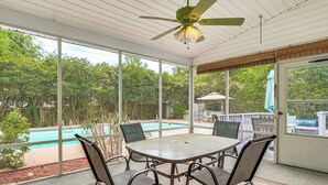 Un patio couvert avec une table et des chaises, un ventilateur de plafond et une vue sur une piscine et des arbres.