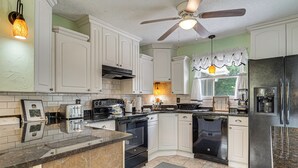 Une cuisine avec des meubles blancs, un réfrigérateur noir, un ventilateur de plafond et une fenêtre avec un rideau.
