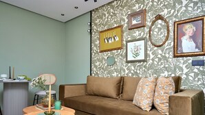 Una habitación con un sofá marrón, una mesa redonda con un florero y libros, un taburete pequeño y una pared con cuadros y un papel pintado estampado.