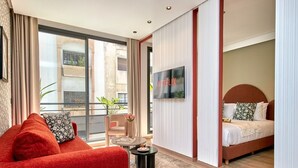 Habitación de hotel con sofá, cama, televisión, balcón con plantas y vista a la ciudad.