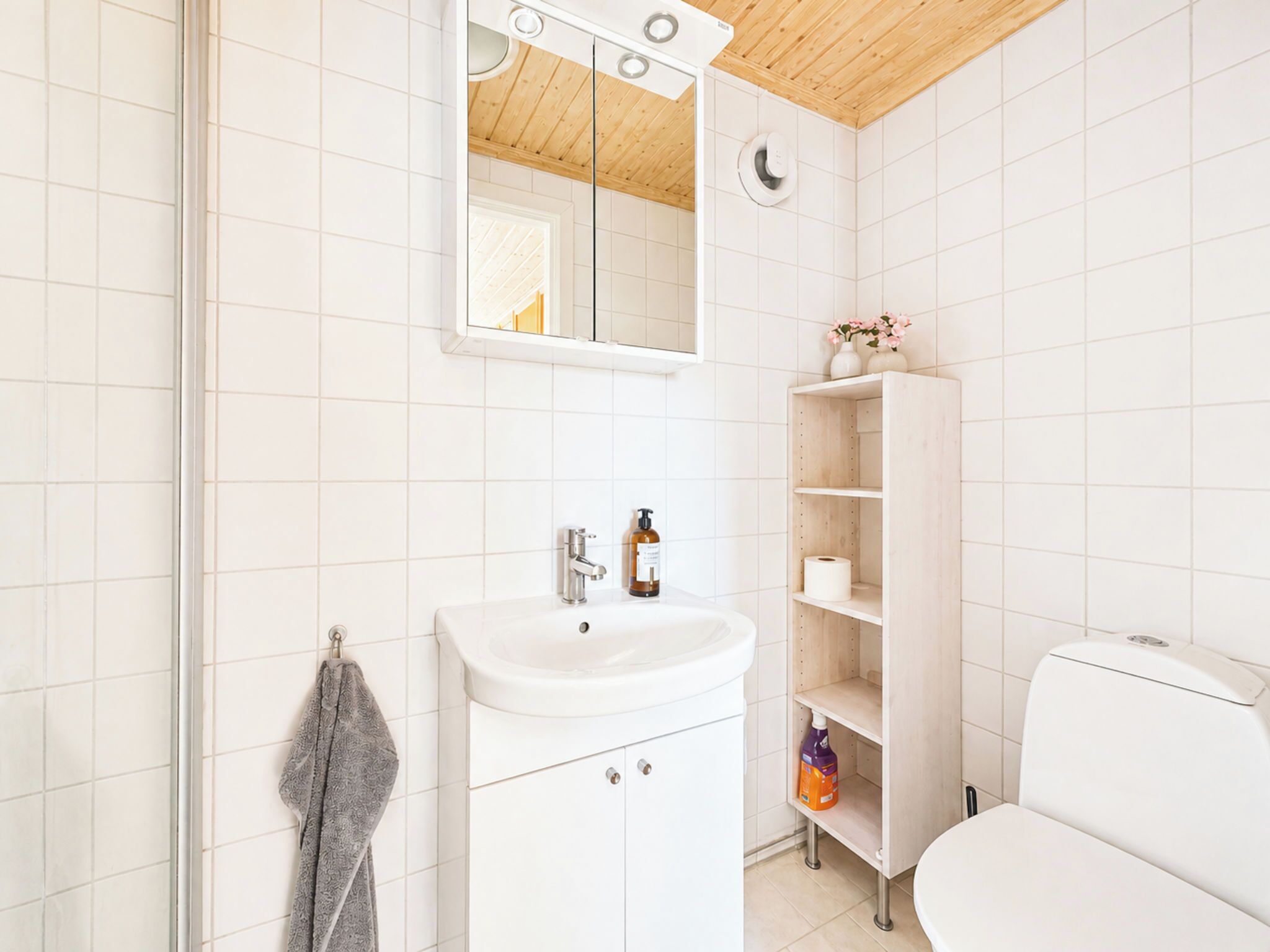 A bathroom with white tiles, a wooden ceiling, a white sink, a mirror, a toilet, and a shelf with toiletries.