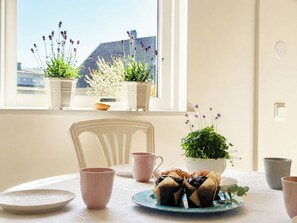 A dining area with a table set for two, a window with potted plants, and a view of buildings outside. - Skeaberg by Interhome (Halmstad)