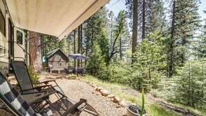 Un espacio al aire libre techado con una silla y una mesa, una cabaña de madera al fondo y un paisaje boscoso.