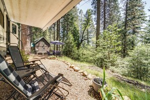 Ein überdachter Außenbereich mit Stuhl und Tisch, im Hintergrund eine Holzhütte und eine bewaldete Landschaft.
