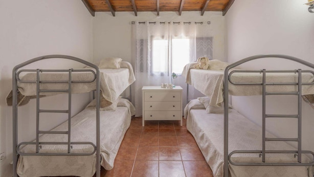 Ein Zimmer mit zwei Etagenbetten, einem kleinen Nachttisch, einem Fenster mit Vorhängen und einer Holzbalkendecke.