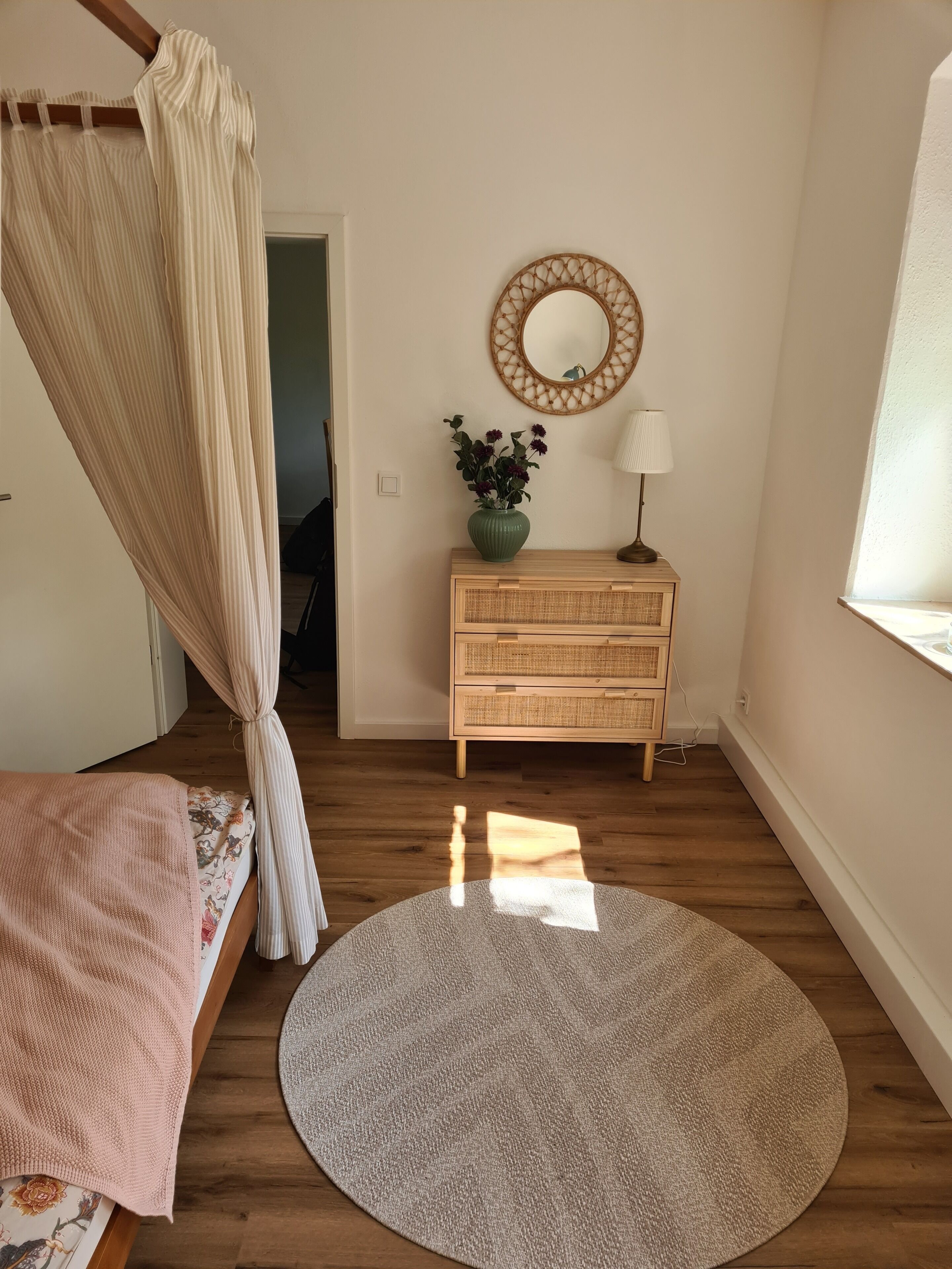 Une chambre à coucher avec un lit, un tapis rond, une commode, un miroir et une fenêtre.