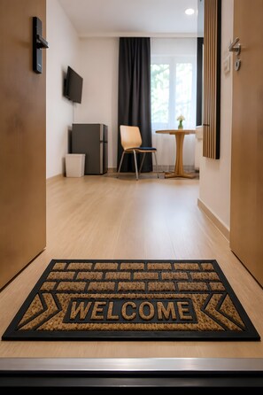 Ein Hotel-Empfangsbereich mit einer 'WELCOME'-Fußmatte, einem Fernseher, einem Sessel, einem kleinen Tisch und einem Fenster mit Vorhängen.
