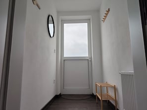 Ein enger Flur mit einer weißen Tür, einem kleinen Holzregal und einer runden Matte auf dem Boden.