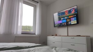 Quarto com cama, janela com cortinas, TV fixada na parede exibindo diversos serviços de streaming e cômoda branca.
