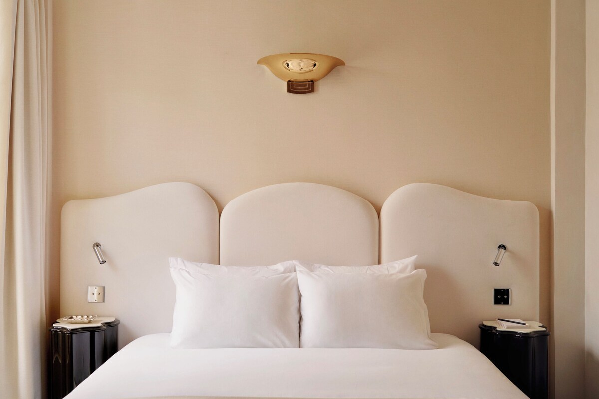 A hotel room with a large bed, a headboard with a light fixture, two bedside tables, and a curtain on the left.