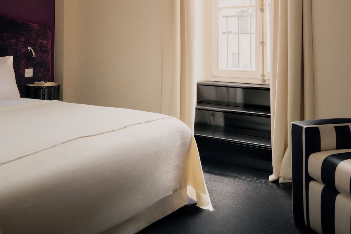 A hotel room with a bed, a window with curtains, a chair, and a piano.