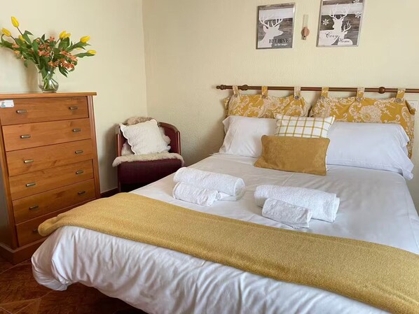 A neatly made bed with white and yellow linens, a wooden dresser with multiple drawers, a chair with a white cushion, and two wall art pieces.