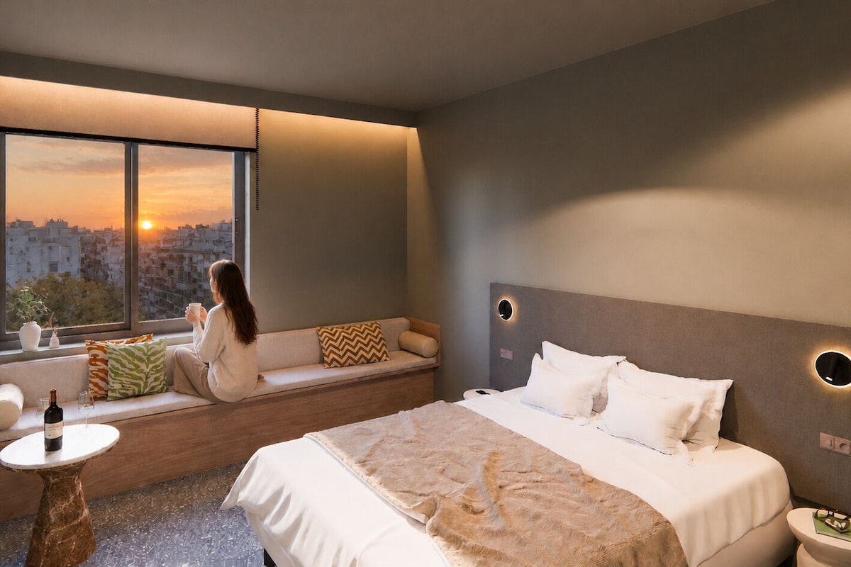 A modern hotel room with a large bed, a seating area by the window, a small table with a bottle and glass, and a nightstand with a lamp.