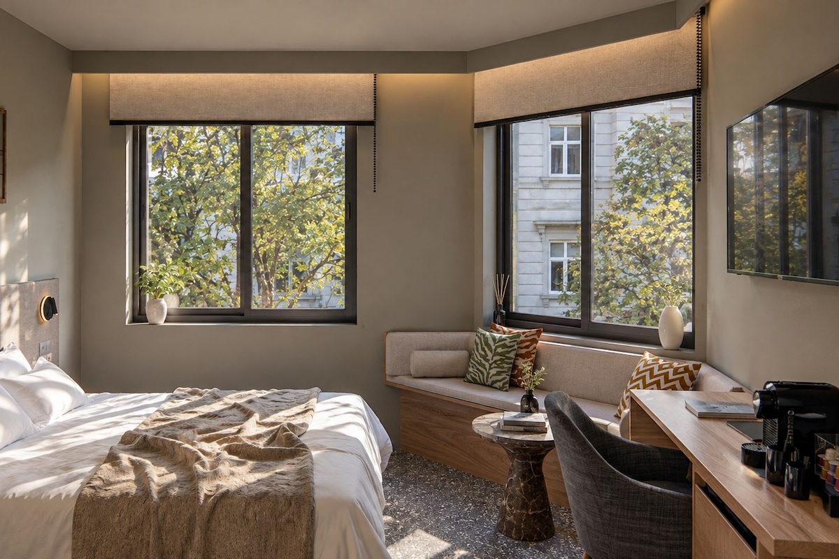 A modern hotel room with a bed, a seating area with a sofa and a table, a desk with a chair, and large windows offering a view of trees and buildings.
