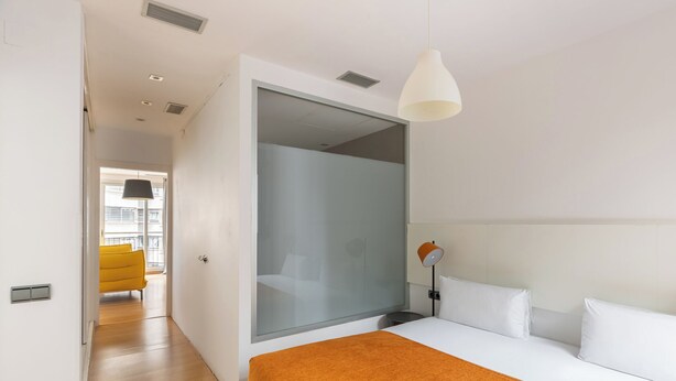 Un hotel moderno con una cama grande, un separador de vidrio, una silla amarillo brillante y vistas a la ciudad.
