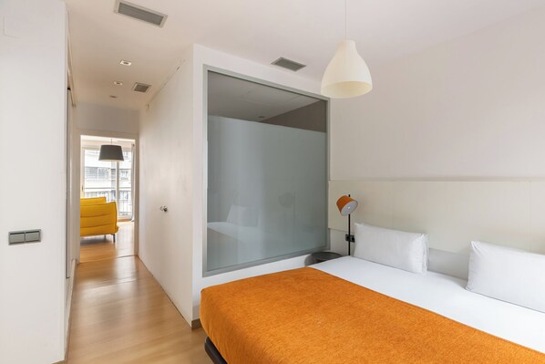 A modern hotel room with a large bed, a glass partition, a bright yellow chair, and a view of the city.
