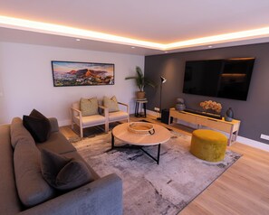 Ein modernes Wohnzimmer mit einer Couch, Sesseln, einem Couchtisch, einem Fernseher und einem Konsolentisch mit Dekoration.