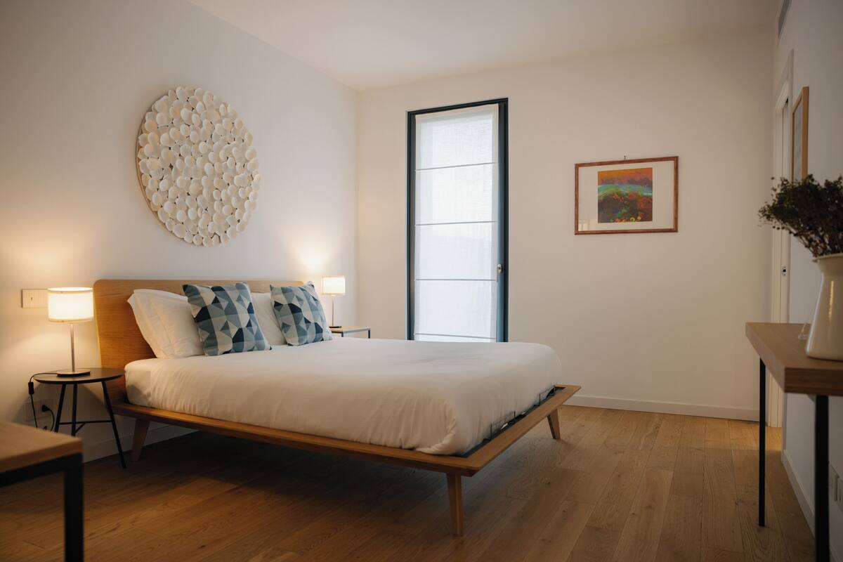 Ein Schlafzimmer mit Bett, Nachttischen, einer Wandleuchte, einem Fenster und einem gerahmten Bild.