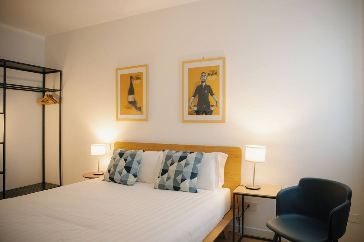 Ein Hotelzimmer mit einem Bett, zwei Nachttischlampen, einem Sessel, einem Kleiderständer und zwei gerahmten Bildern an der Wand.