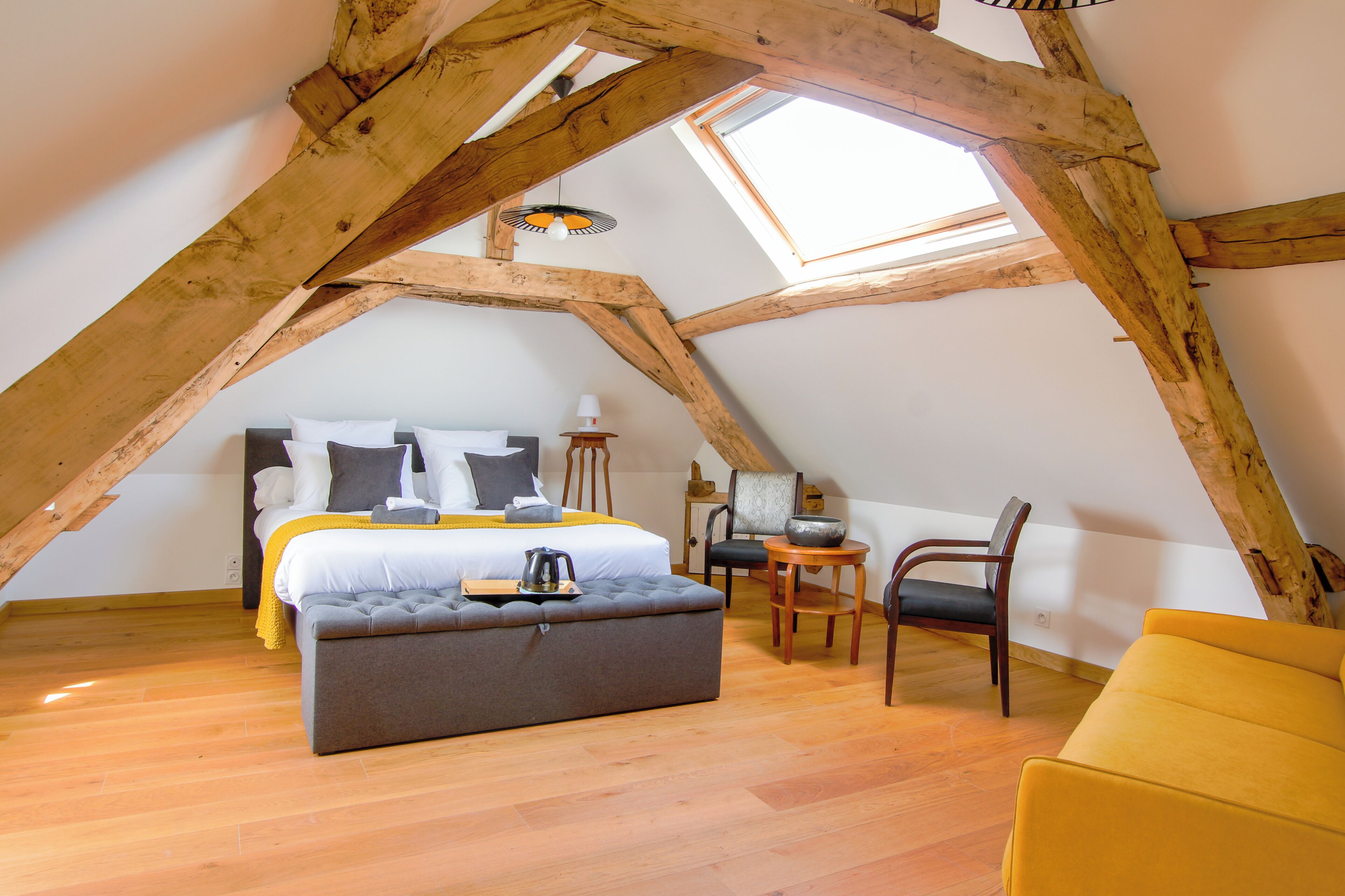 A loft bedroom with a sloped ceiling, a bed with pillows, a bedside table, a chair, and a small table with a lamp.