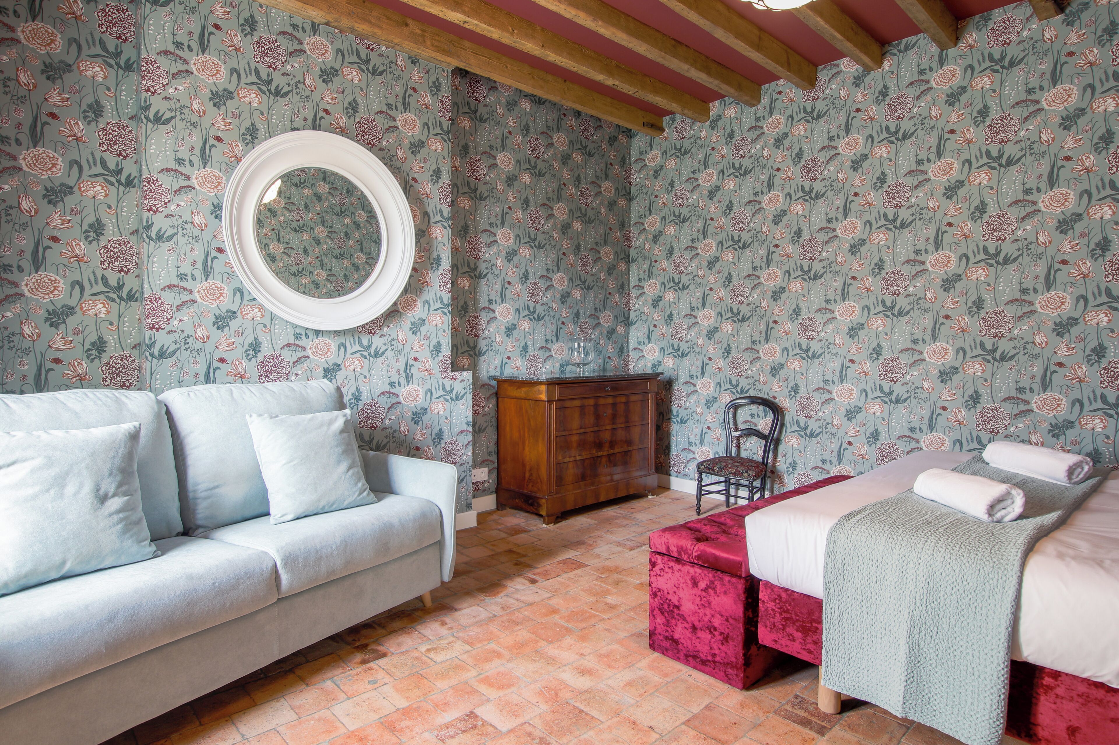 Un dormitorio con papel tapiz floral, un espejo redondo, un aparador de madera, un reposapiés rojo, una cama con una colcha verde y un sofá blanco.