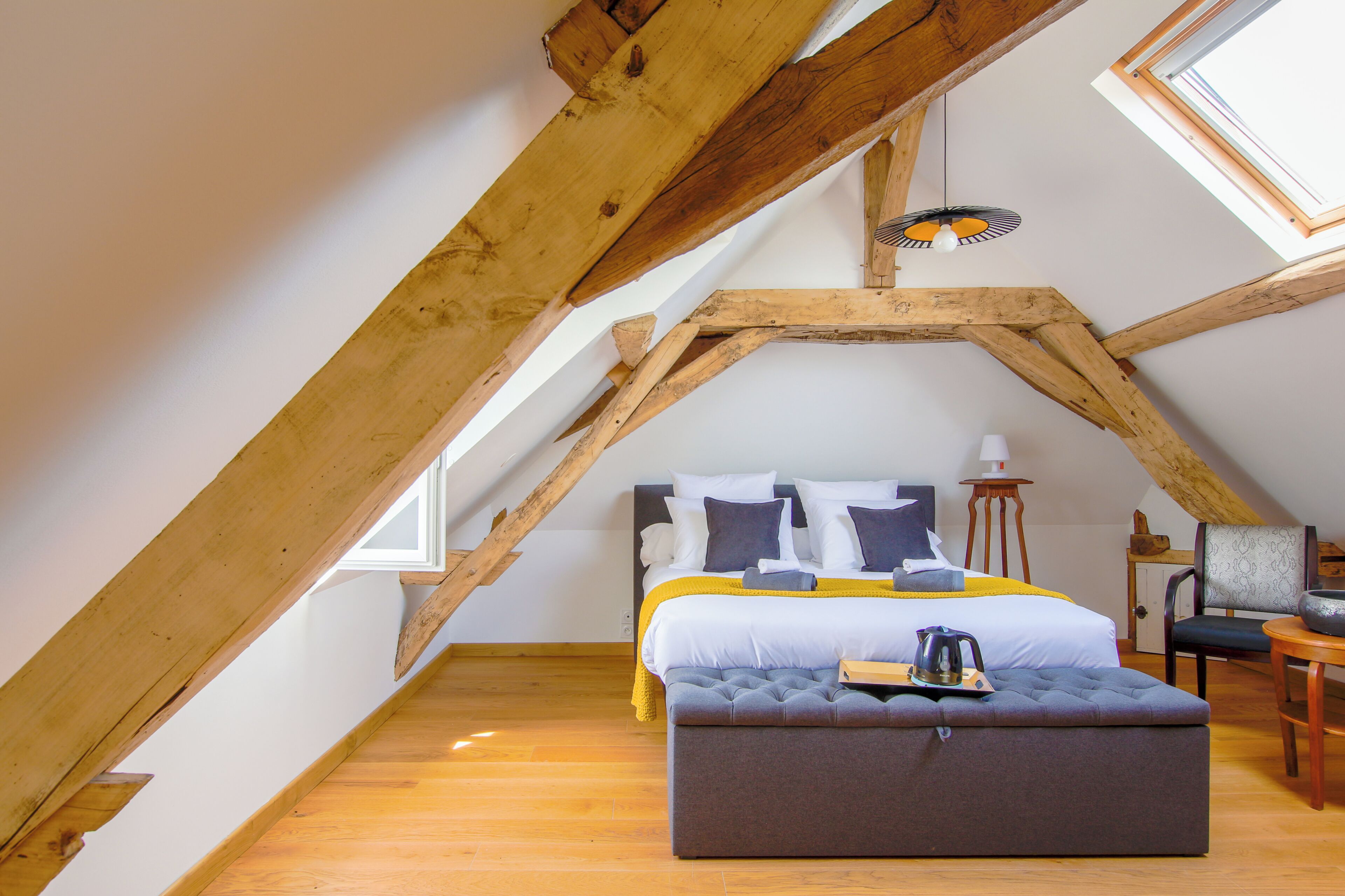 A bedroom with a sloped ceiling, a bed with pillows, a bedside table, a chair, and a small table with a kettle.