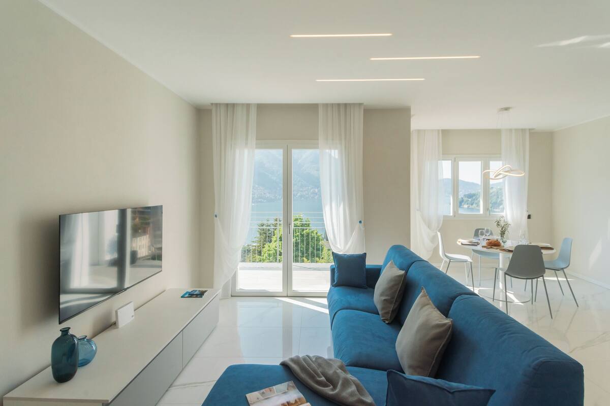 A modern living room with a flat-screen TV, a sofa, a dining area, and a view of the outdoors.