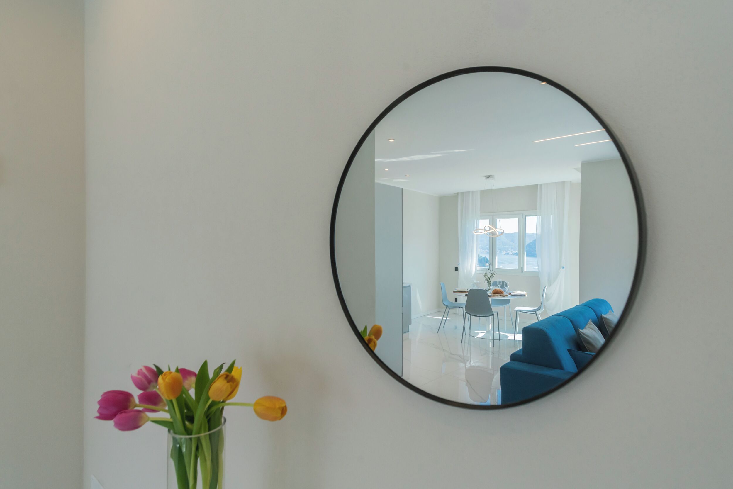 A round mirror on a wall, reflecting a room with a blue sofa, a dining table, and chairs.