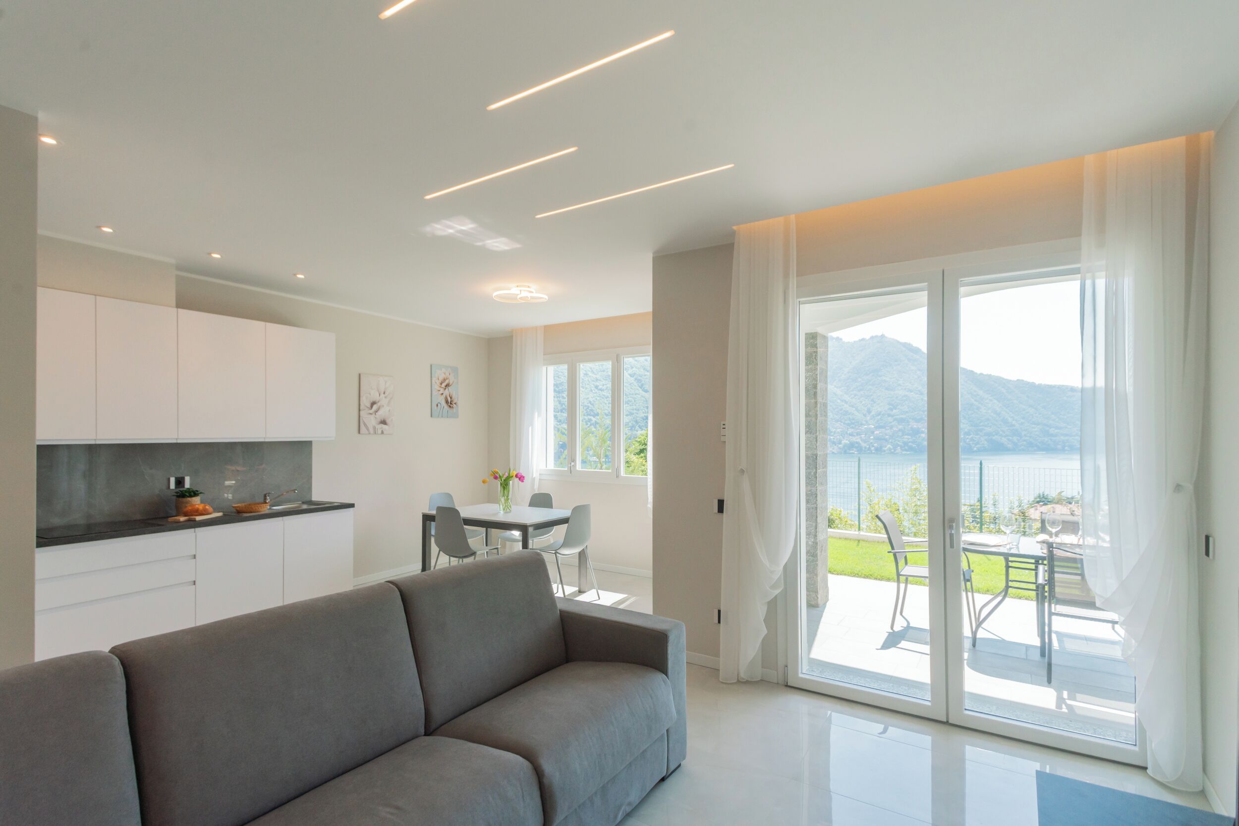 A modern living room with a sofa, a dining area with chairs, a kitchen with cabinets, and a large sliding glass door leading to a balcony with a view of mountains.