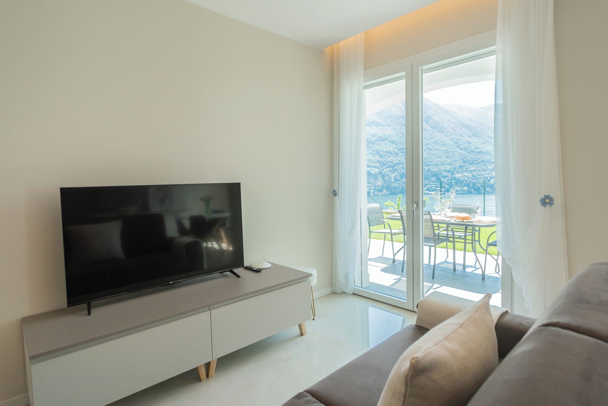 A modern living room with a flat-screen TV on a white cabinet, a sofa, and a sliding glass door leading to an outdoor area with a table and chairs.