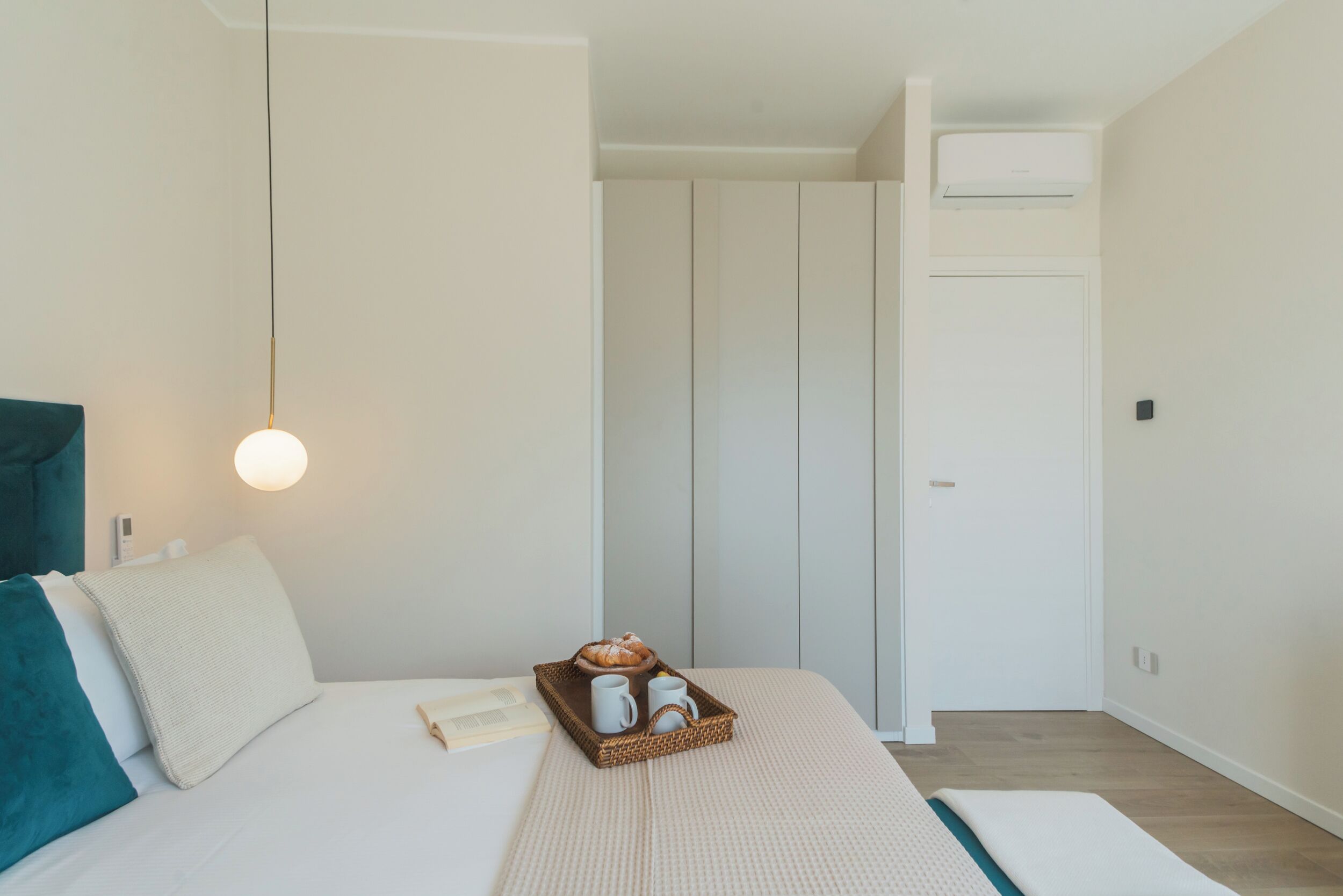A bedroom with a bed, a bedside table, a tray with a croissant, and a wall-mounted air conditioner.