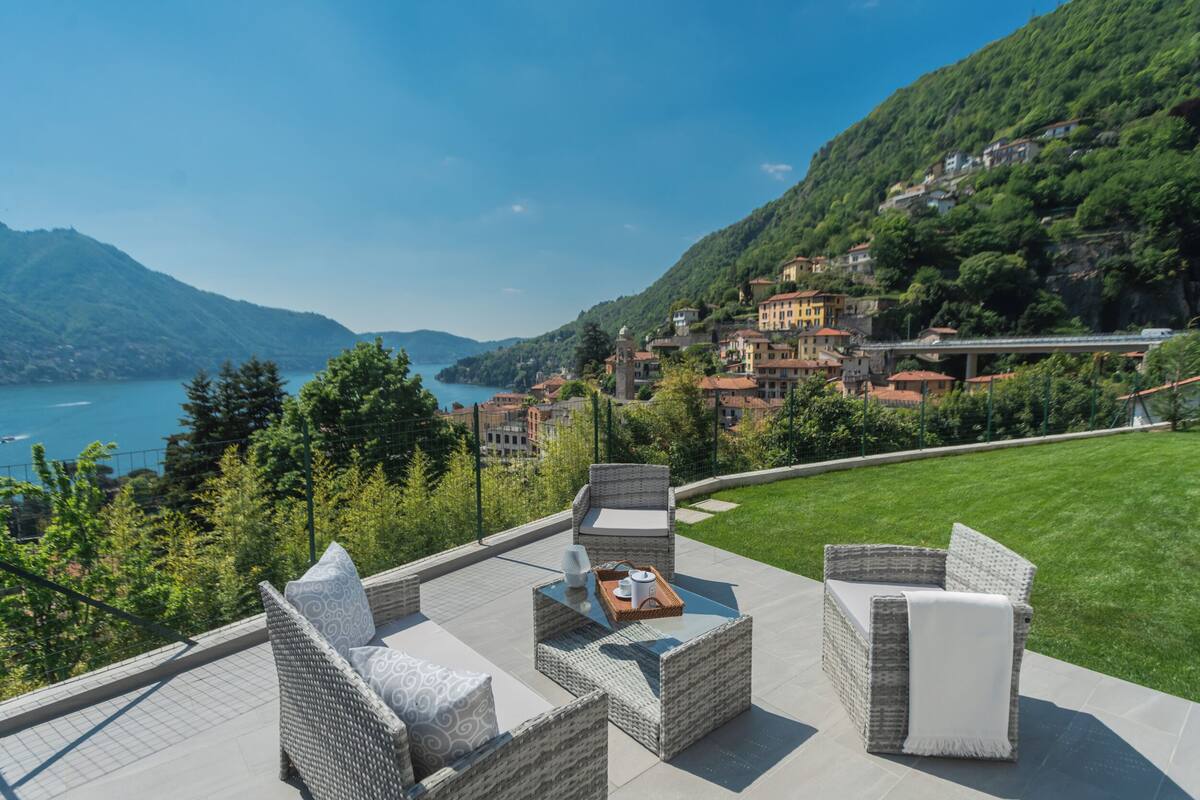 A terrace with wicker furniture, a glass table, and a view of a lake and buildings.