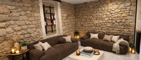 A cozy living room with stone walls, two brown sofas, a beige ottoman, and a bookshelf.