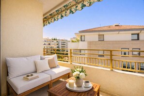 A balcony with a white sofa, a wooden table with a tray, a vase of flowers, and a railing. - Odyssey Love Suite - Croisette - Double Bathtub (Cannes)