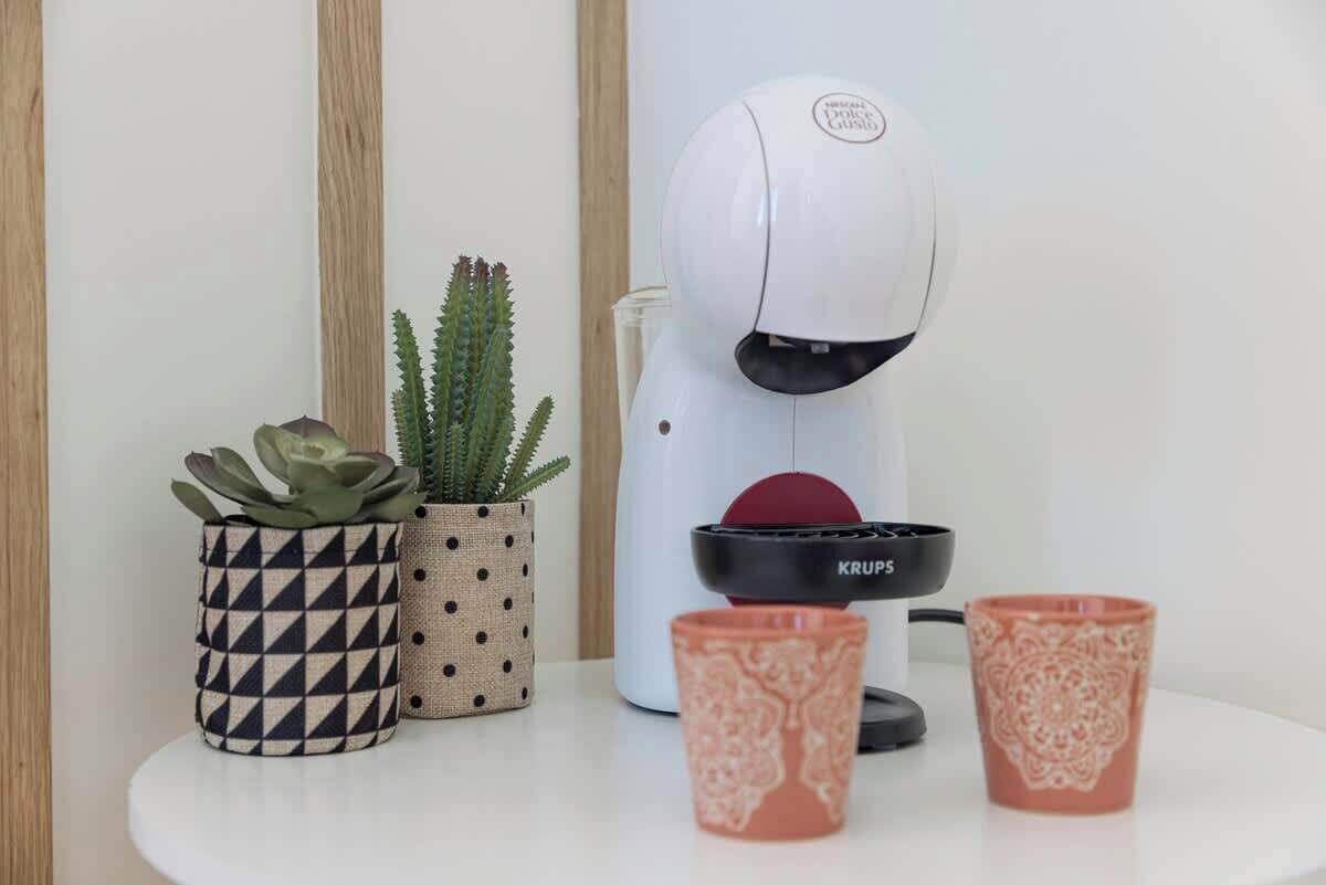 Eine weiße Kaffeemaschine, zwei gemusterte Tassen und zwei Topfpflanzen auf einem weißen Tisch.