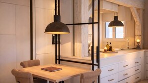 Un comedor con mesa y sillas de madera, una lámpara colgante, y una cocina con armarios blancos y un ventanal.
