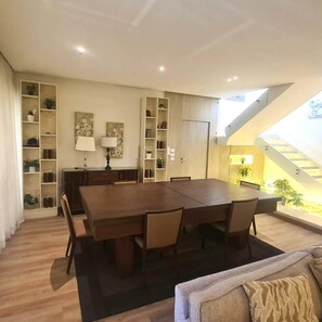 A dining area with a large table and chairs, surrounded by shelves with plants and books, and a staircase leading to an upper level. - Breeze @ Lakeview Res I Nesty Hospitality (New Cairo City)