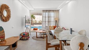 Una habitación moderna con una mesa de vidrio, sillas de madera, un sofá, una alfombra pequeña, una lámpara y vistas a una piscina.