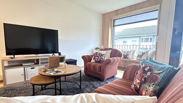 A living room with a TV, sofa, armchair, coffee table, and a window with a view of buildings. - "AllgäuLiebe", Oberstaufen (Oberstaufen)