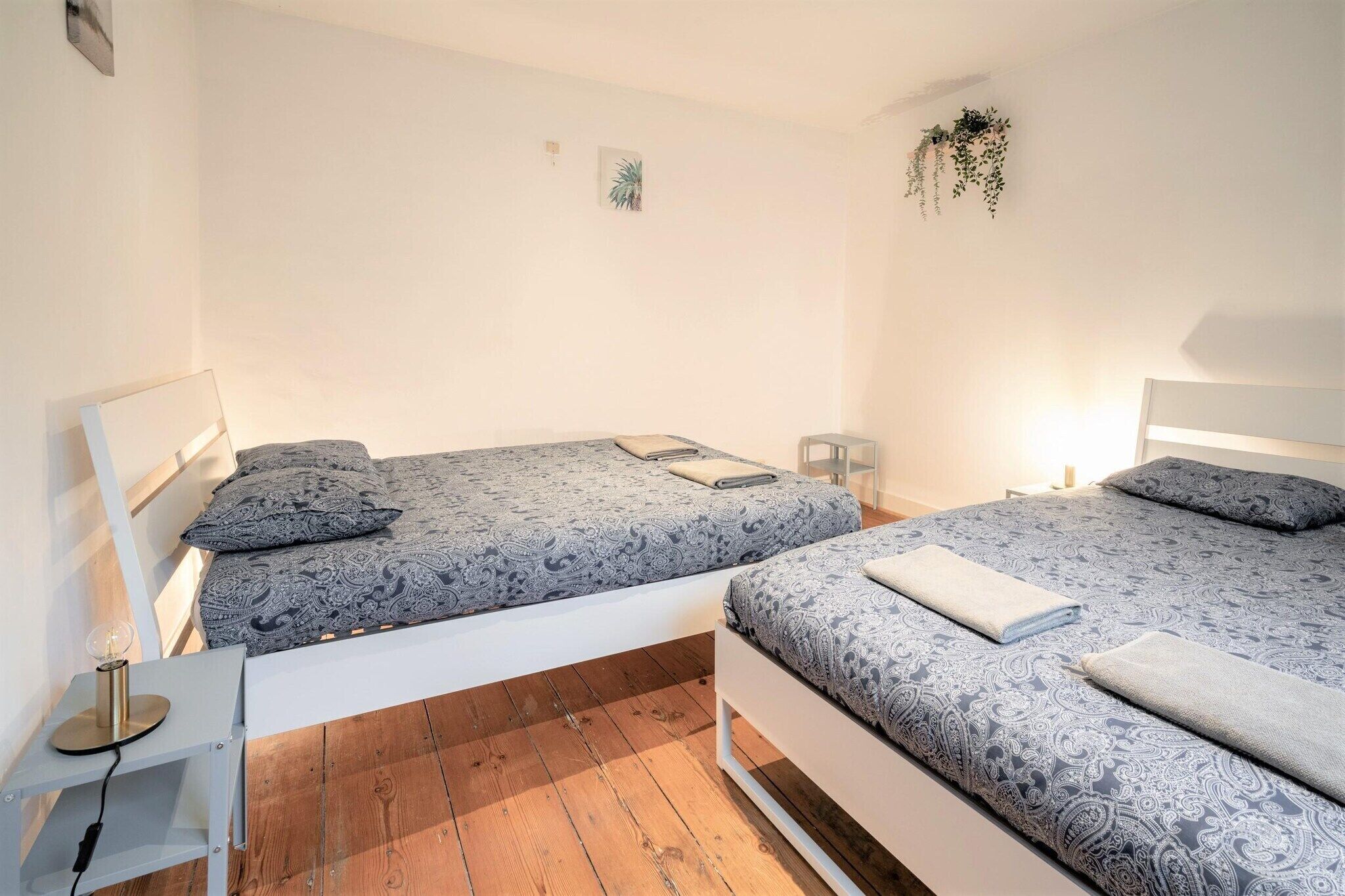 Camera matrimoniale con due letti, un comodino con lampada, una piccola pianta e un punto luce a parete.