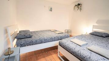 Camera matrimoniale con due letti, un comodino con lampada, una piccola pianta e un punto luce a parete.