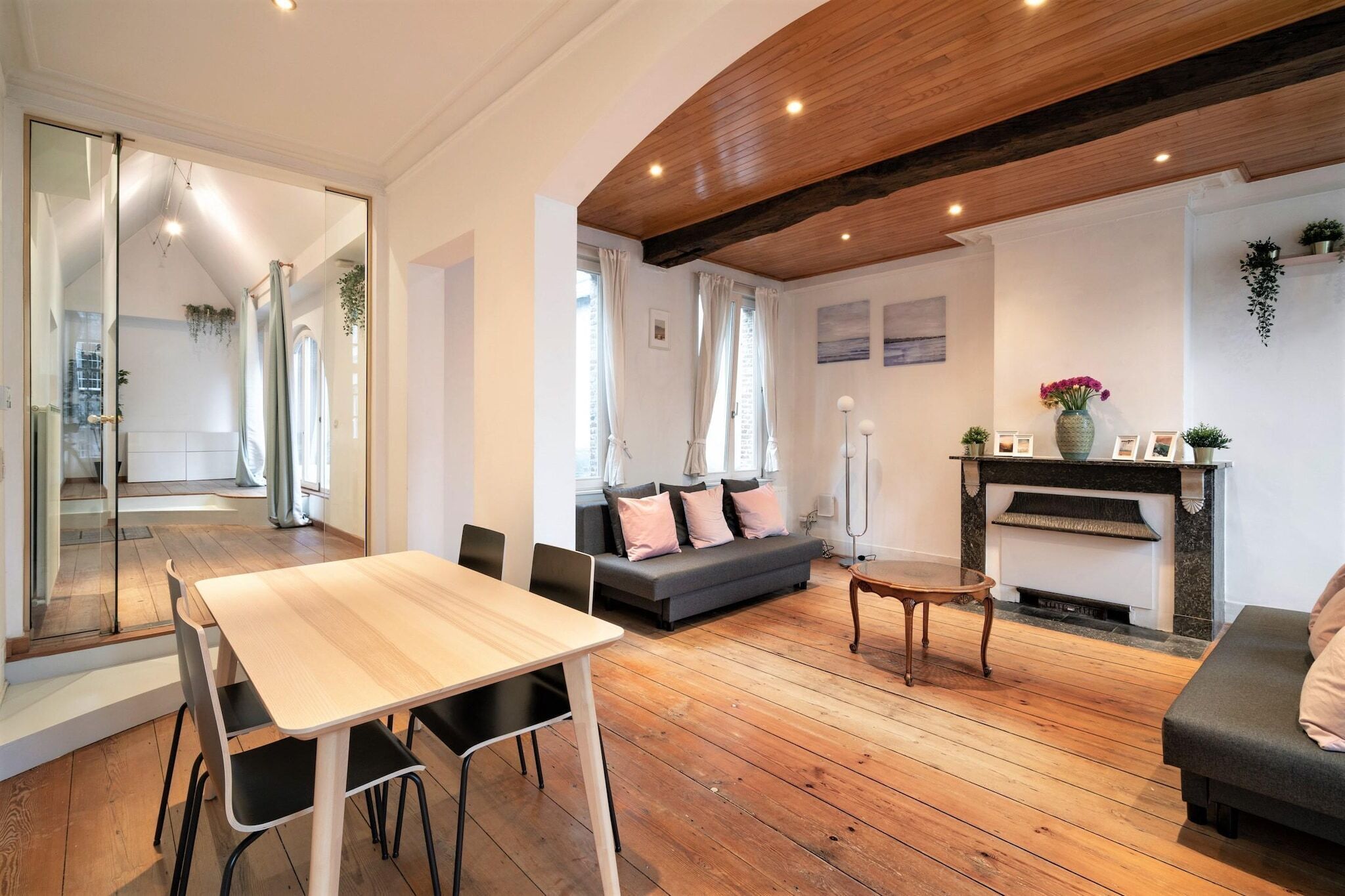 A modern living room with a wooden floor, a sofa, a dining table with chairs, a fireplace, and a large mirror.