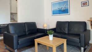 A living room with two black leather sofas, a wooden coffee table, a small potted plant, a framed painting of a sailboat, and a kitchen area with white cabinets and a stove.